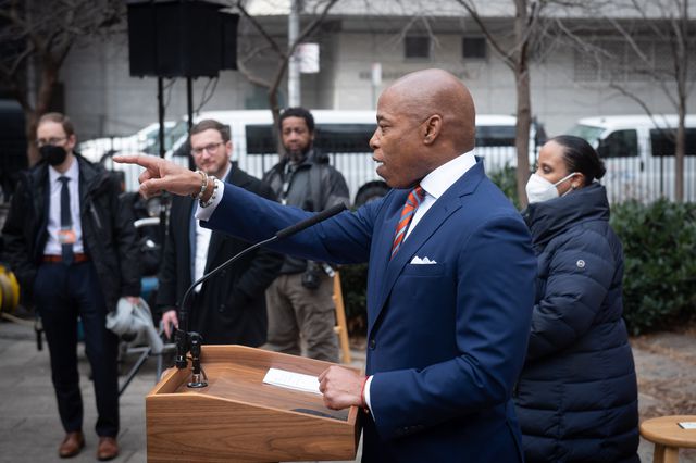 Mayor Eric Adams stands in front of a podium fielding questions from reporters on January 13, 2022. In the background are several officials in Adams' administration.